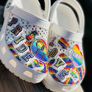 May include: White clogs decorated with rainbow-colored rhinestones and patches. The patches include the words "Pride", "Love is Love", and "Proud!" along with a rainbow heart and a peace sign.