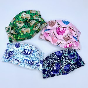 Puede incluir: Cuatro mascarillas de tela con estampado de perezoso. Las mascarillas son de color verde, rosa, azul y blanco con diferentes diseños de perezosos.