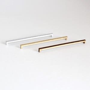 May include: Three linear cabinet pulls arranged diagonally, featuring a slim rectangular profile. The set includes one silver-toned pull, one gold-toned pull, and one dark bronze-toned pull, each with a smooth finish and two small mounting posts.