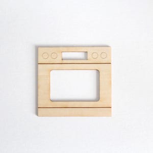 May include: A flat, light-colored wooden cutout shaped like the front panel of an oven. The piece features a rectangular cutout for the oven door, a horizontal control panel area with four circular knob outlines, and a central rectangular opening.