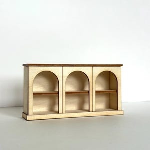 May include: A small wooden bookshelf featuring three arched compartments and shelves. Constructed from light-toned wood, it has darker wood detailing on the top and shelves. This miniature bookshelf is designed with a simple, elegant style, ideal for dollhouses or small displays.