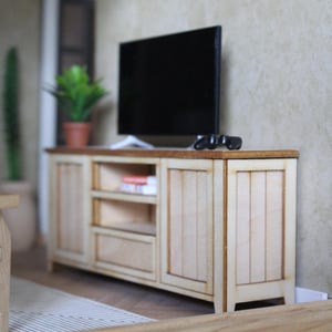 May include: Miniature wooden TV cabinet with a dark brown top and light wood doors and drawers. A black television sits on top, with a small controller. A potted plant and a cactus are in the background.