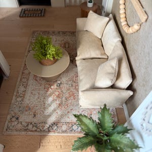 K&ouml;nnte beinhalten: Eine Miniatur-Wohnzimmerszene mit einem beigen Sofa und passenden Kissen, einem runden Couchtisch mit einer gr&uuml;nen Pflanze, einem gemusterten Teppich in Orange- und Graut&ouml;nen, einem Wandbehang aus Holzperlen und einem kleinen Kaktus im Topf.
