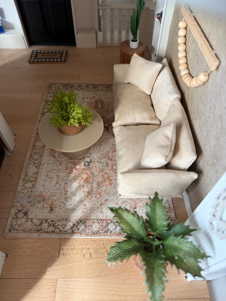K&ouml;nnte beinhalten: Eine Miniatur-Wohnzimmerszene mit einem beigen Sofa und passenden Kissen, einem runden Couchtisch mit einer gr&uuml;nen Pflanze, einem gemusterten Teppich in Orange- und Graut&ouml;nen, einem Wandbehang aus Holzperlen und einem kleinen Kaktus im Topf.
