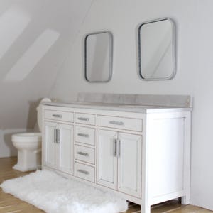 May include: A miniature bathroom vanity in white with a marble-look countertop, silver-toned hardware, and four central drawers flanked by two cabinets. Two rectangular mirrors with silver frames are mounted above, with a white toilet nearby.