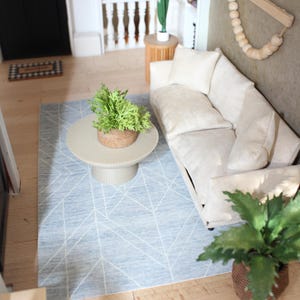 K&ouml;nnte beinhalten: Eine Miniatur-Wohnzimmerszene mit einem cremefarbenen Sofa und passenden Kissen, einem runden beigen Couchtisch mit einer gr&uuml;nen Pflanze, einem hellblauen Teppich mit wei&szlig;en geometrischen Linien und einem Wandbehang aus Holz mit Perlen.