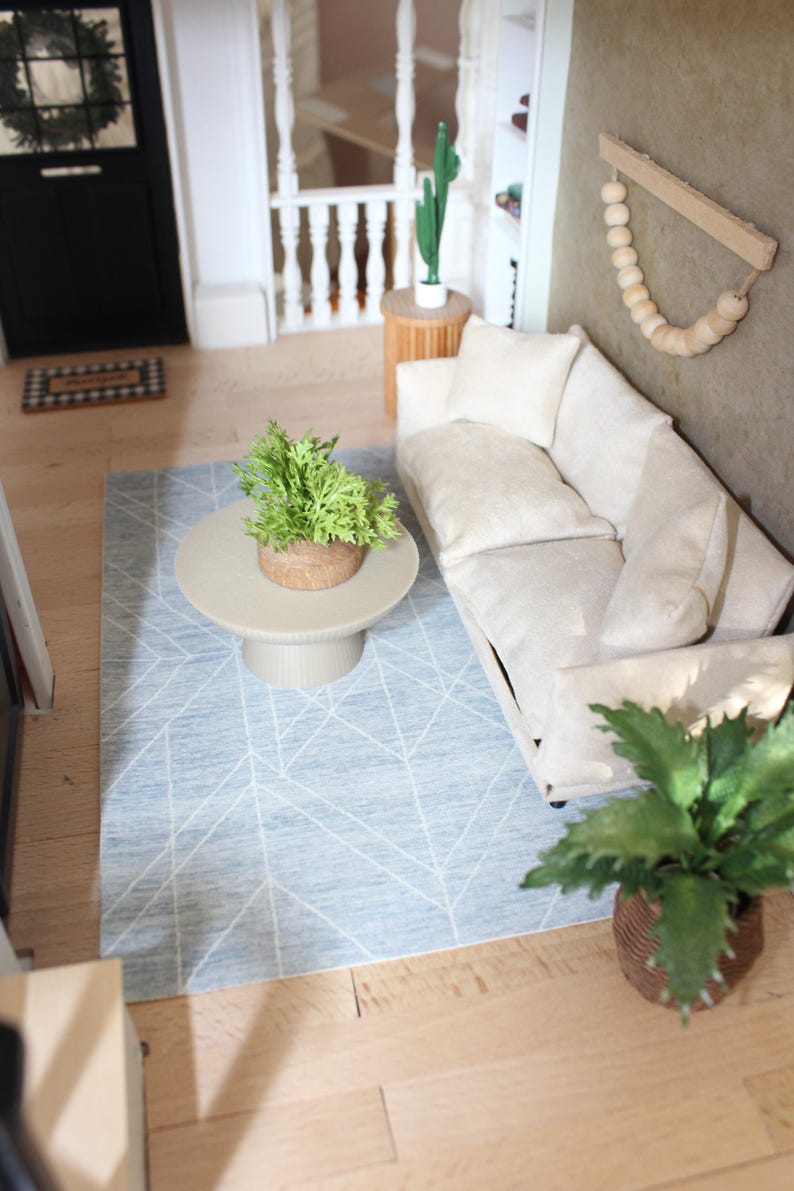 K&ouml;nnte beinhalten: Eine Miniatur-Wohnzimmerszene mit einem cremefarbenen Sofa und passenden Kissen, einem runden beigen Couchtisch mit einer gr&uuml;nen Pflanze, einem hellblauen Teppich mit wei&szlig;en geometrischen Linien und einem Wandbehang aus Holz mit Perlen.