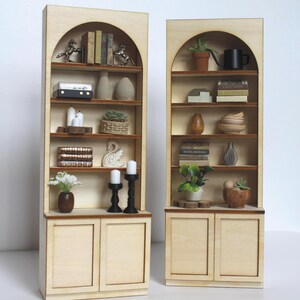 May include: Two miniature wooden bookshelves with arched tops. Each shelf displays small decorative items like books, vases, candles, and plants. The shelves are light-coloured with a natural wood grain, and the doors at the bottom are also made of wood.