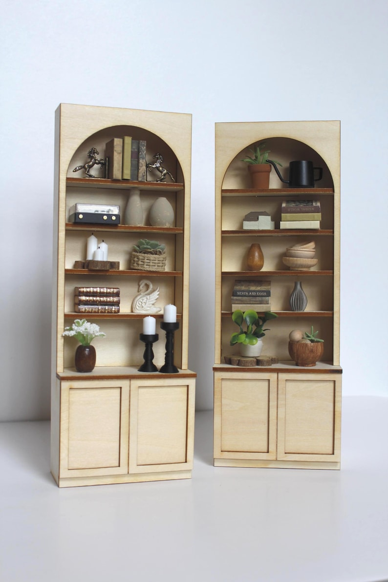 May include: Two miniature wooden bookshelves with arched tops. Each shelf displays small decorative items like books, vases, candles, and plants. The shelves are light-coloured with a natural wood grain, and the doors at the bottom are also made of wood.