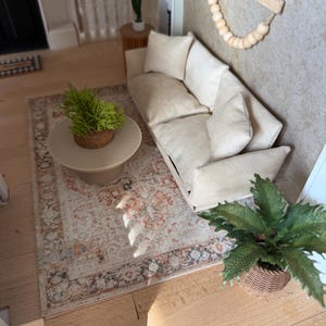 K&ouml;nnte beinhalten: Eine Miniatur-Wohnzimmerszene mit einem beigen Sofa und passenden Kissen, einem runden beigen Couchtisch mit einer kleinen gr&uuml;nen Pflanze, einem gemusterten Teppich in Orange- und Graut&ouml;nen sowie einem Wandbehang aus Holzperlen.