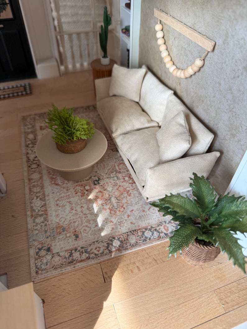 K&ouml;nnte beinhalten: Eine Miniatur-Wohnzimmerszene mit einem beigen Sofa und passenden Kissen, einem runden beigen Couchtisch mit einer kleinen gr&uuml;nen Pflanze, einem gemusterten Teppich in Orange- und Graut&ouml;nen sowie einem Wandbehang aus Holzperlen.