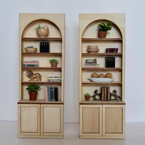 May include: Two miniature wooden bookshelves with arched tops. Each shelf displays small decorative items like potted plants, books, bowls, and figurines. The shelves are made of light-colored wood and have a simple, elegant design.