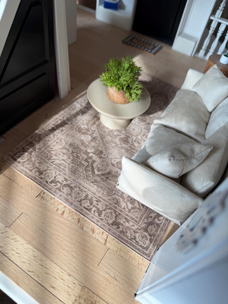 K&ouml;nnte beinhalten: Eine Miniatur-Puppenhaus-Wohnzimmerszene mit einem cremefarbenen Sofa inklusive passender Kissen, einem runden, hellen Couchtisch mit S&auml;ulenfu&szlig; und einer kleinen gr&uuml;nen Topfpflanze. Darunter liegt ein rechteckiger Teppich mit braun-cremefarbenem Muster.