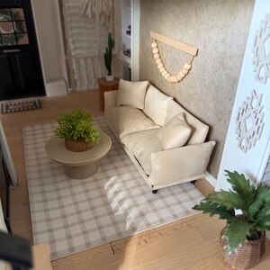K&ouml;nnte beinhalten: Eine Miniatur-Wohnzimmerszene mit einem beigen Sofa mit passenden Zierkissen, einem runden beigen Couchtisch mit einer gr&uuml;nen Pflanze und einem beigewei&szlig;en karierten Teppich. Ein Wandbehang aus Holzperlen ist an der strukturierten beigen Wand befestigt.