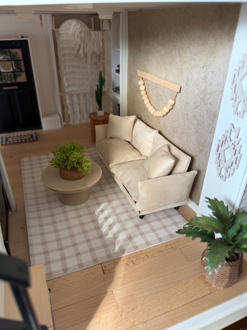 K&ouml;nnte beinhalten: Eine Miniatur-Wohnzimmerszene mit einem beigen Sofa mit passenden Zierkissen, einem runden beigen Couchtisch mit einer gr&uuml;nen Pflanze und einem beigewei&szlig;en karierten Teppich. Ein Wandbehang aus Holzperlen ist an der strukturierten beigen Wand befestigt.