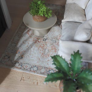 K&ouml;nnte beinhalten: Eine Miniatur-Wohnzimmerszene mit einem cremefarbenen Sofa mit weichen Kissen, einem runden beigen Couchtisch mit Sockel und einer kleinen gr&uuml;nen Topfpflanze sowie einem rechteckigen Teppich mit verblasstem orange-blau-cremefarbenem Muster.