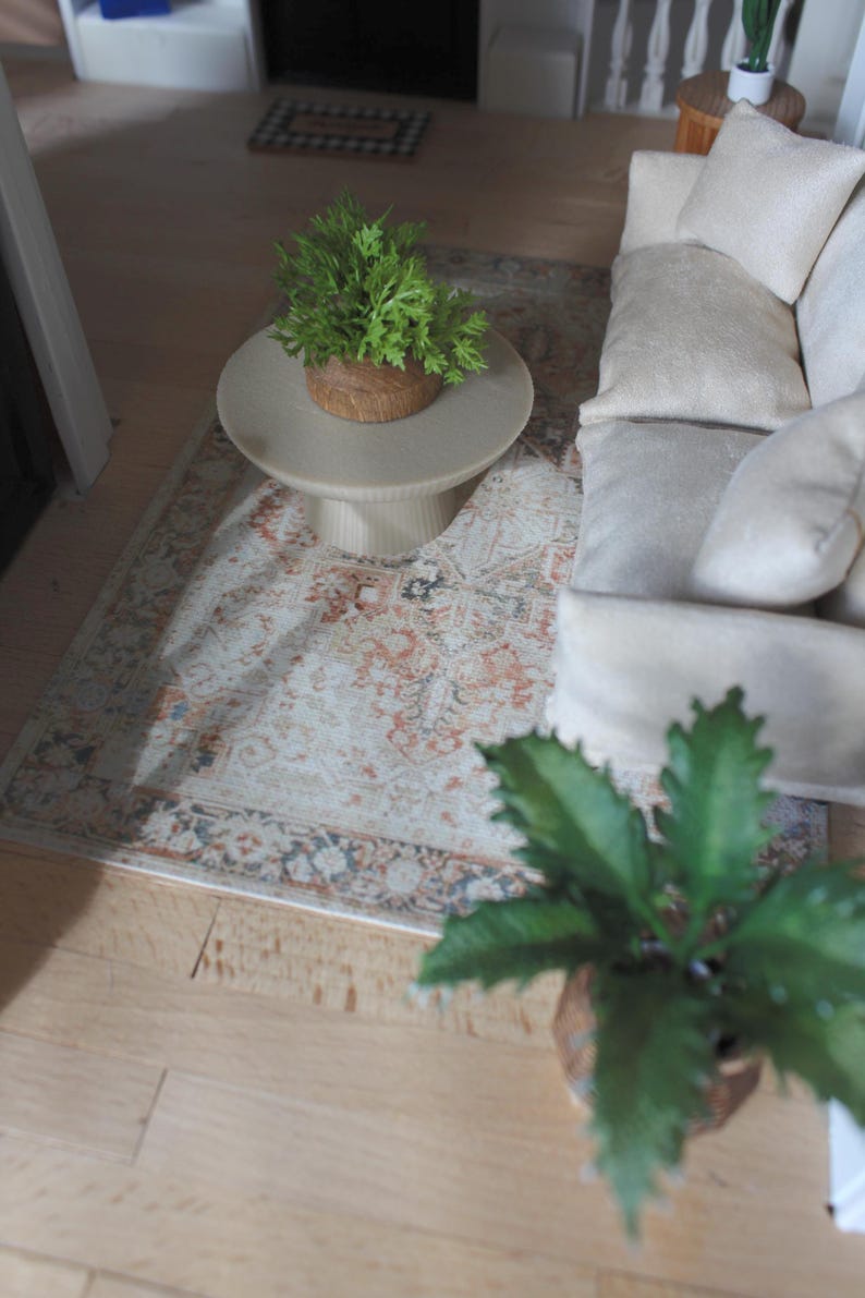 K&ouml;nnte beinhalten: Eine Miniatur-Wohnzimmerszene mit einem cremefarbenen Sofa mit weichen Kissen, einem runden beigen Couchtisch mit Sockel und einer kleinen gr&uuml;nen Topfpflanze sowie einem rechteckigen Teppich mit verblasstem orange-blau-cremefarbenem Muster.