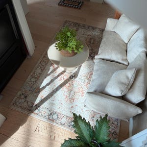 K&ouml;nnte beinhalten: Ein Miniatur-Wohnzimmer-Set f&uuml;r Puppenh&auml;user mit einem cremefarbenen, strukturierten Sofa inklusive passender Kissen, einem runden beigen Couchtisch mit Sockel und einem gemusterten Teppich in Orange-, Blau- und Cremet&ouml;nen sowie zwei gr&uuml;nen Topfpflanzen.