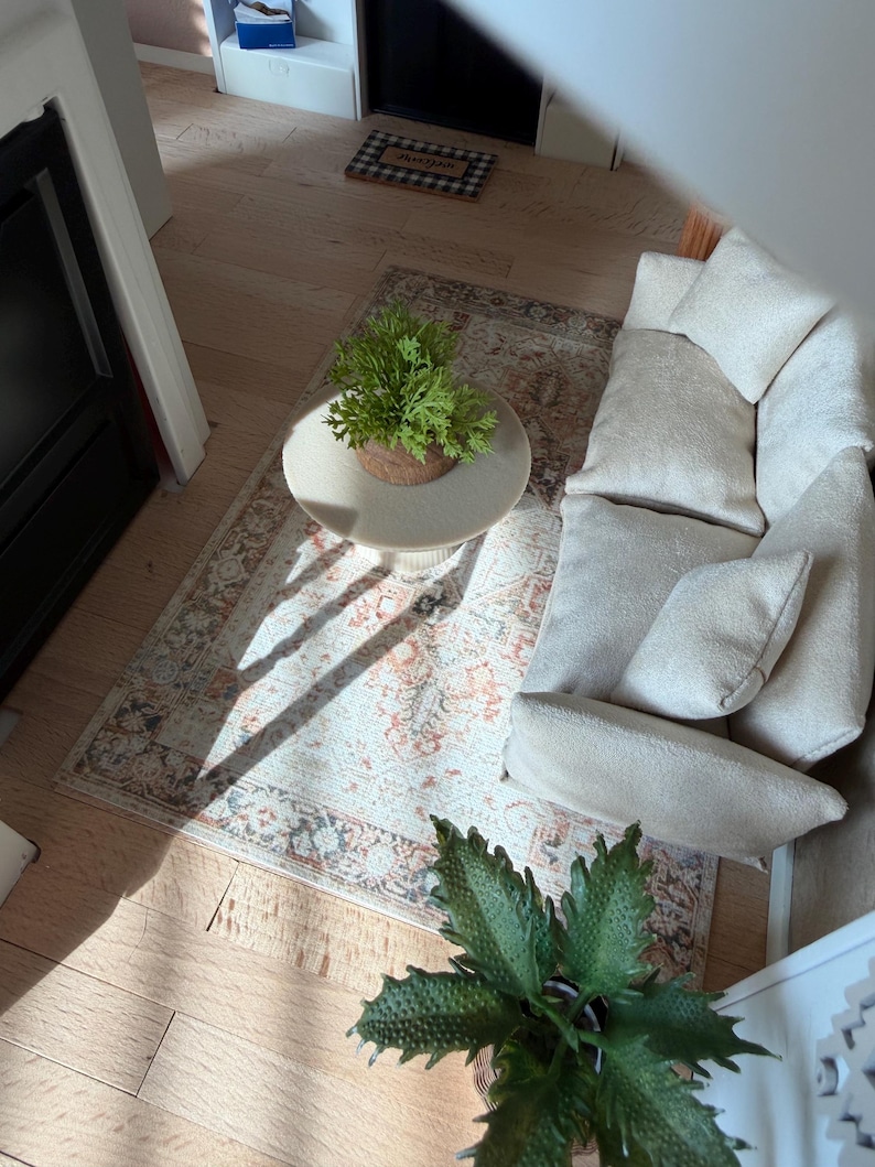 K&ouml;nnte beinhalten: Ein Miniatur-Wohnzimmer-Set f&uuml;r Puppenh&auml;user mit einem cremefarbenen, strukturierten Sofa inklusive passender Kissen, einem runden beigen Couchtisch mit Sockel und einem gemusterten Teppich in Orange-, Blau- und Cremet&ouml;nen sowie zwei gr&uuml;nen Topfpflanzen.