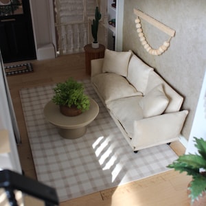 K&ouml;nnte beinhalten: Ein Miniatur-Puppenhaus-Wohnzimmer mit einem beigen Sofa und zwei passenden Kissen, einem runden beigen Couchtisch mit einer gr&uuml;nen Topfpflanze, einem beigewei&szlig;en karierten Teppich und einem Wandbehang aus Holzperlen an einer beigen Wand.