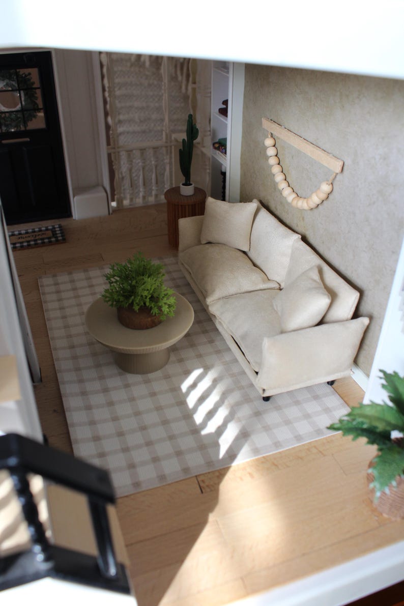 K&ouml;nnte beinhalten: Ein Miniatur-Puppenhaus-Wohnzimmer mit einem beigen Sofa und zwei passenden Kissen, einem runden beigen Couchtisch mit einer gr&uuml;nen Topfpflanze, einem beigewei&szlig;en karierten Teppich und einem Wandbehang aus Holzperlen an einer beigen Wand.