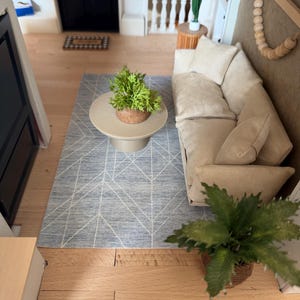 K&ouml;nnte beinhalten: Ein Miniatur-Wohnzimmer-Set bestehend aus einem beigen Sofa mit passenden Kissen, einem runden beigen Couchtisch mit einer kleinen gr&uuml;nen Pflanze und einem blauen Teppich mit wei&szlig;em geometrischem Linienmuster. Eine Holzperlengirlande h&auml;ngt an der Wand.