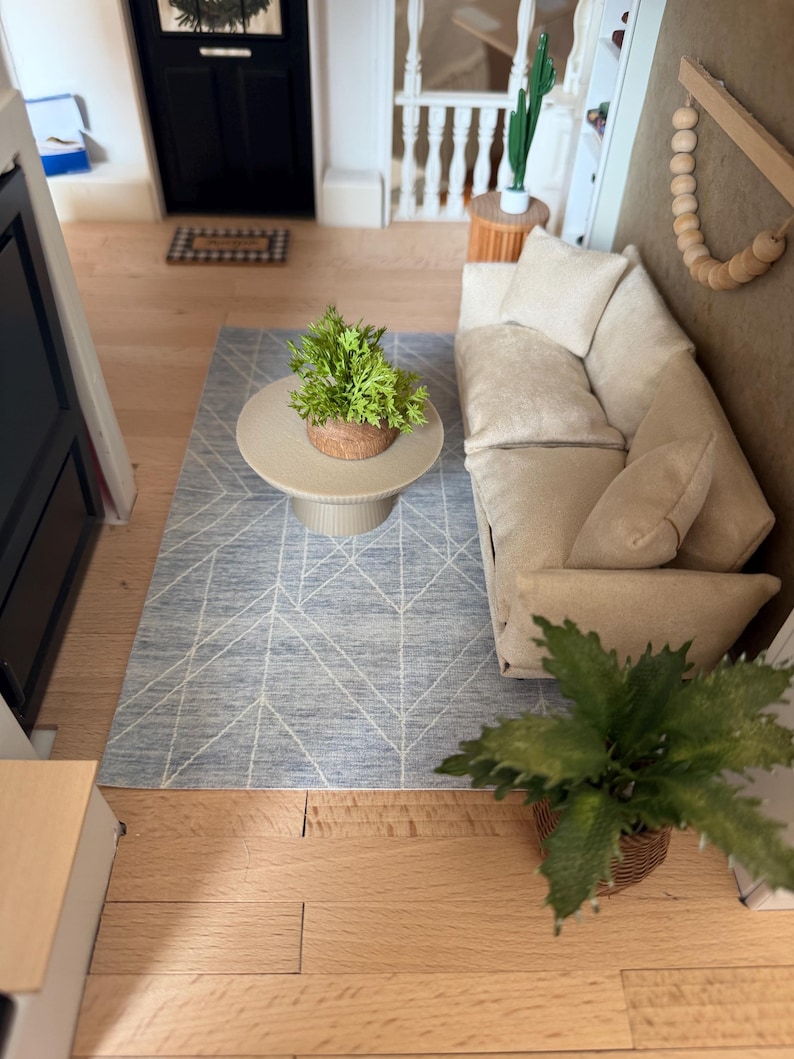 K&ouml;nnte beinhalten: Ein Miniatur-Wohnzimmer-Set bestehend aus einem beigen Sofa mit passenden Kissen, einem runden beigen Couchtisch mit einer kleinen gr&uuml;nen Pflanze und einem blauen Teppich mit wei&szlig;em geometrischem Linienmuster. Eine Holzperlengirlande h&auml;ngt an der Wand.