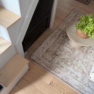 K&ouml;nnte beinhalten: Eine Miniatur-Puppenhaustreppe mit wei&szlig;en Setzstufen und hellen Holzstufen, platziert neben einem kleinen beigen Teppich mit Muster. Ein winziger runder Beistelltisch tr&auml;gt eine kleine gr&uuml;ne Topfpflanze mit einem braunen Sockel.