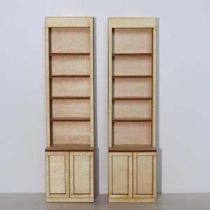May include: Two miniature wooden bookshelves with a light wood finish. Each bookshelf has four shelves and a two-door cabinet at the base. The shelves and cabinet doors are a darker shade of brown. The background is plain white.