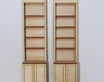 May include: Two miniature wooden bookshelves with a light wood finish. Each bookshelf has four shelves and a two-door cabinet at the base. The shelves and cabinet doors are a darker shade of brown. The background is plain white.