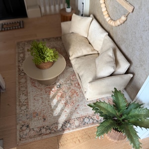 K&ouml;nnte beinhalten: Eine Miniatur-Wohnzimmerszene mit einem beigen Sofa und passenden Kissen, einem runden beigen Couchtisch mit einer gr&uuml;nen Pflanze, einem gemusterten Teppich in Orange- und Graut&ouml;nen sowie einem Wandbehang aus Holzperlen.