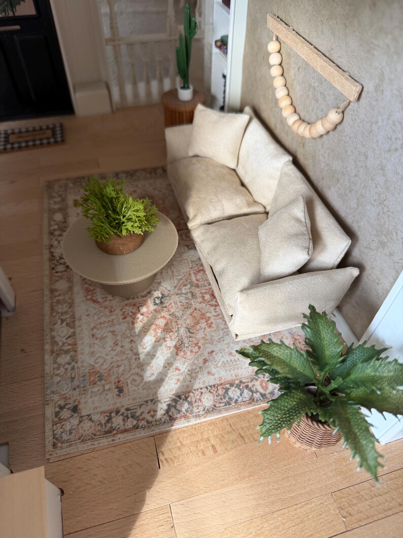 K&ouml;nnte beinhalten: Eine Miniatur-Wohnzimmerszene mit einem beigen Sofa und passenden Kissen, einem runden beigen Couchtisch mit einer gr&uuml;nen Pflanze, einem gemusterten Teppich in Orange- und Graut&ouml;nen sowie einem Wandbehang aus Holzperlen.