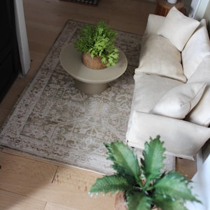 K&ouml;nnte beinhalten: Eine Miniatur-Wohnzimmerszene mit einem cremefarbenen Sofa mit passenden Zierkissen, einem runden beigen Couchtisch mit Sockel und einem rechteckigen Teppich mit verblasstem beige-wei&szlig;em Muster. Zwei kleine gr&uuml;ne Topfpflanzen sind enthalten.