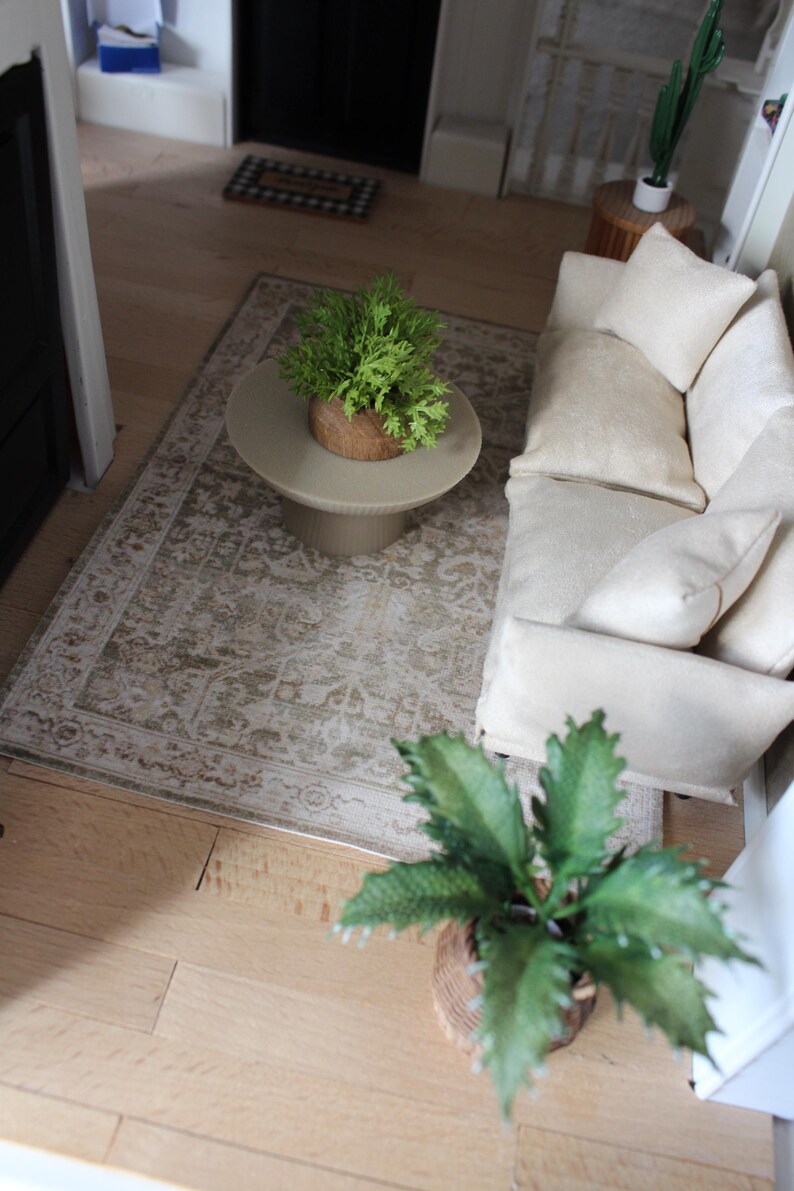 K&ouml;nnte beinhalten: Eine Miniatur-Wohnzimmerszene mit einem cremefarbenen Sofa mit passenden Zierkissen, einem runden beigen Couchtisch mit Sockel und einem rechteckigen Teppich mit verblasstem beige-wei&szlig;em Muster. Zwei kleine gr&uuml;ne Topfpflanzen sind enthalten.