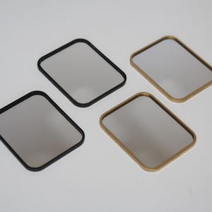 May include: Four rectangular mirrors with rounded corners. Two mirrors have black frames, and two have gold frames. The mirror surfaces are a light grey colour. The mirrors are arranged on a white surface.