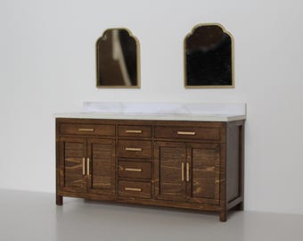 May include: A miniature bathroom vanity with a dark wood finish, featuring multiple drawers and cabinet doors. It has a white countertop and is paired with two arched mirrors with gold frames. The overall design is modern.
