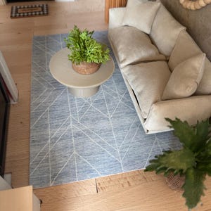 K&ouml;nnte beinhalten: Ein Miniatur-Wohnzimmerset bestehend aus einem beigen Sofa mit drei Kissen, einem runden beigen Couchtisch mit geriffeltem Fu&szlig; und einem blauen Teppich mit wei&szlig;em geometrischem Linienmuster. Eine kleine gr&uuml;ne Pflanze steht auf dem Couchtisch.