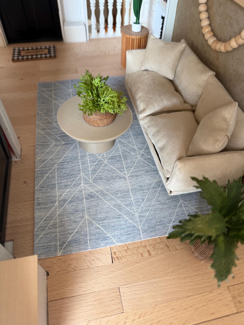 K&ouml;nnte beinhalten: Ein Miniatur-Wohnzimmerset bestehend aus einem beigen Sofa mit drei Kissen, einem runden beigen Couchtisch mit geriffeltem Fu&szlig; und einem blauen Teppich mit wei&szlig;em geometrischem Linienmuster. Eine kleine gr&uuml;ne Pflanze steht auf dem Couchtisch.