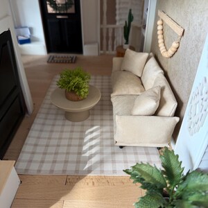 K&ouml;nnte beinhalten: Eine Miniatur-Wohnzimmerszene mit einem beigen Sofa samt passenden Kissen, einem runden Couchtisch auf einem Sockel mit einer gr&uuml;nen Topfpflanze und einem hellen karierten Teppich. Ein Wandbehang aus Holzperlen und eine Pflanze im Korb.