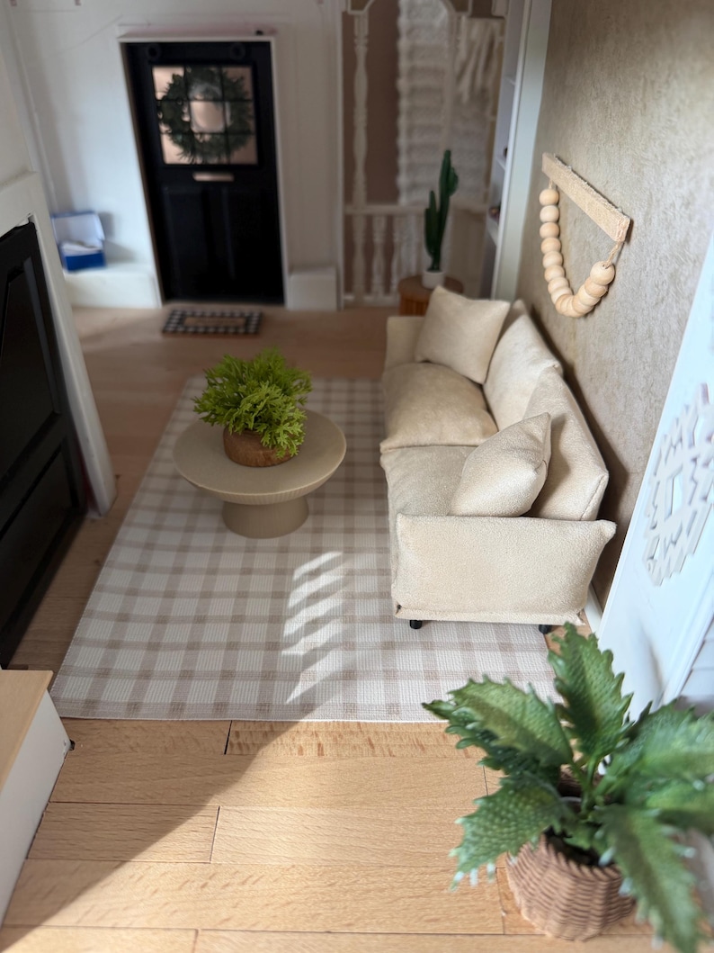K&ouml;nnte beinhalten: Eine Miniatur-Wohnzimmerszene mit einem beigen Sofa samt passenden Kissen, einem runden Couchtisch auf einem Sockel mit einer gr&uuml;nen Topfpflanze und einem hellen karierten Teppich. Ein Wandbehang aus Holzperlen und eine Pflanze im Korb.