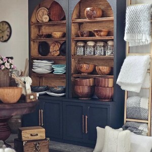 May include: A miniature dark blue cabinet with arched shelves filled with wooden bowls, plates, and jars. The cabinet has a light brown interior and copper-coloured handles. White pillows and blankets are nearby, along with small wooden boxes.