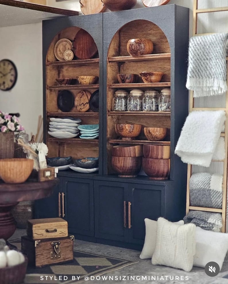May include: A miniature dark blue cabinet with arched shelves filled with wooden bowls, plates, and jars. The cabinet has a light brown interior and copper-coloured handles. White pillows and blankets are nearby, along with small wooden boxes.