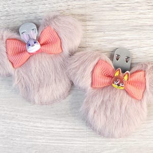 Puede incluir: Dos pinzas para el pelo rosas claras y esponjosas con forma de orejas de rat&oacute;n, cada una con un lazo coral y un dije de personaje. Una tiene un conejo, la otra un zorro. Sobre una superficie de vetas de madera clara.