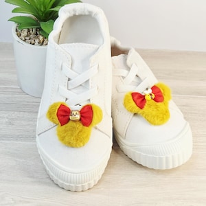May include: A pair of white canvas sneakers with a yellow and red bow embellishment on the toes. The bow has a small gold-colored charm. The shoes have white laces and a white rubber sole.
