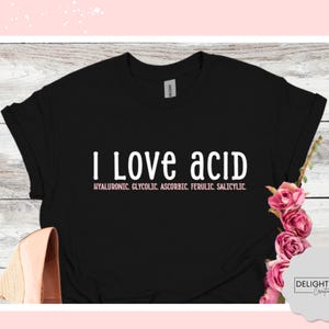 May include: Black t-shirt with the text "I Love acid" in white, with a list of acids below in pink. The shirt is displayed with a pair of shoes and pink flowers.