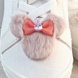 Puede incluir: Primer plano de un zapato de lona blanco con un adorno decorativo. El adorno presenta un lazo rosa con una peque&ntilde;a figura de conejo en el centro y detalles de piel rosa esponjosa. El zapato es de estilo casual.