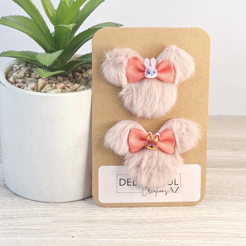 Puede incluir: Dos pinzas para el pelo rosas y esponjosas con forma de cabeza de rat&oacute;n, con lazos color coral y peque&ntilde;os adornos de personajes. Las pinzas est&aacute;n sujetas a una tarjeta marr&oacute;n con el texto "DEL Creations AZ". Una peque&ntilde;a planta en maceta est&aacute; en el fondo.