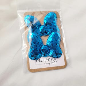 The image displays two shiny, turquoise sequin bows packaged in a clear plastic sleeve. The bows are attached to a cardboard backing with the branding "Delightful Creations AZ" printed at the bottom. The reflective, vibrant sequins give the bows a striking appearance, suggesting their use as decorative accessories. The background is clean and neutral, drawing attention to the product.