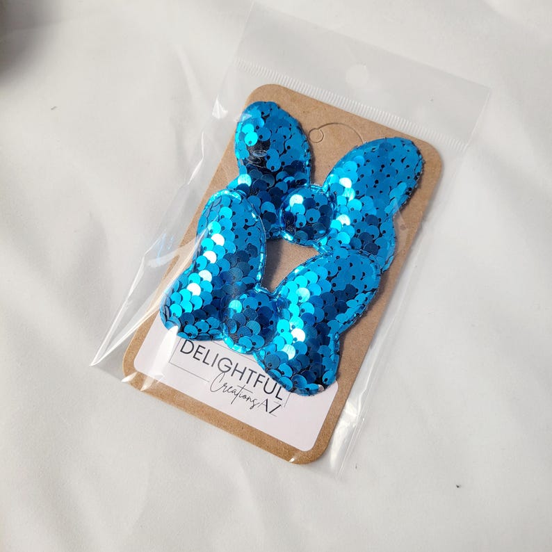 The image displays two shiny, turquoise sequin bows packaged in a clear plastic sleeve. The bows are attached to a cardboard backing with the branding "Delightful Creations AZ" printed at the bottom. The reflective, vibrant sequins give the bows a striking appearance, suggesting their use as decorative accessories. The background is clean and neutral, drawing attention to the product.