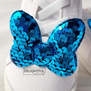 A close-up of a white sneaker with laces, showcasing a Minnie Mouse bow-shaped decoration attached to the front. The bow is adorned with shiny turquoise sequins, creating a vibrant and sparkling appearance. At the bottom of the image, the text "Delightful Creations AZ" emphasizes the branding, adding a personal and professional flair to the accessory.